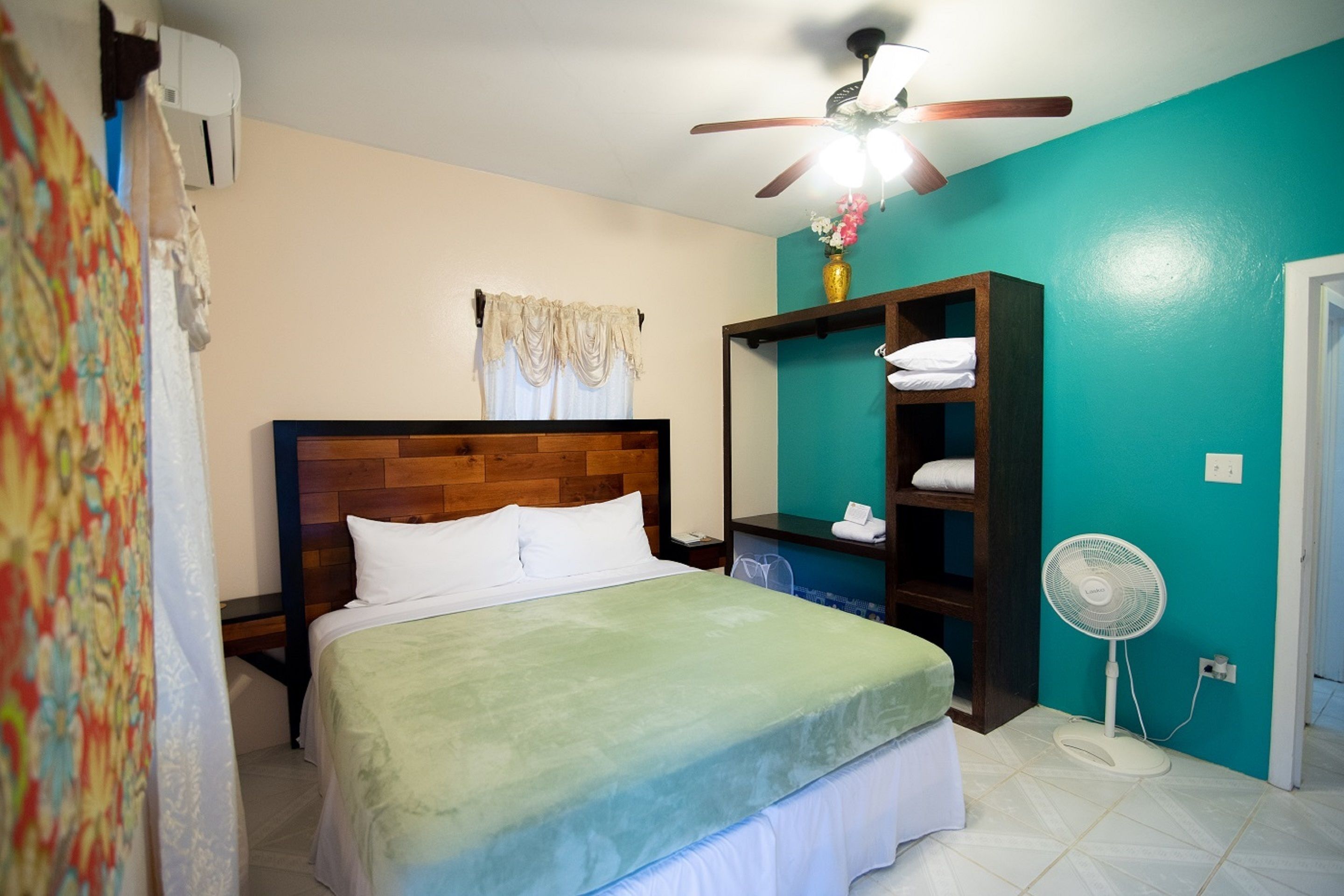 Ein Schlafzimmer mit Holz-Kopfteil, einem Bett mit weißer Bettwäsche, einer türkisblauen Wand, einem Deckenventilator und einem weißen Ventilator.