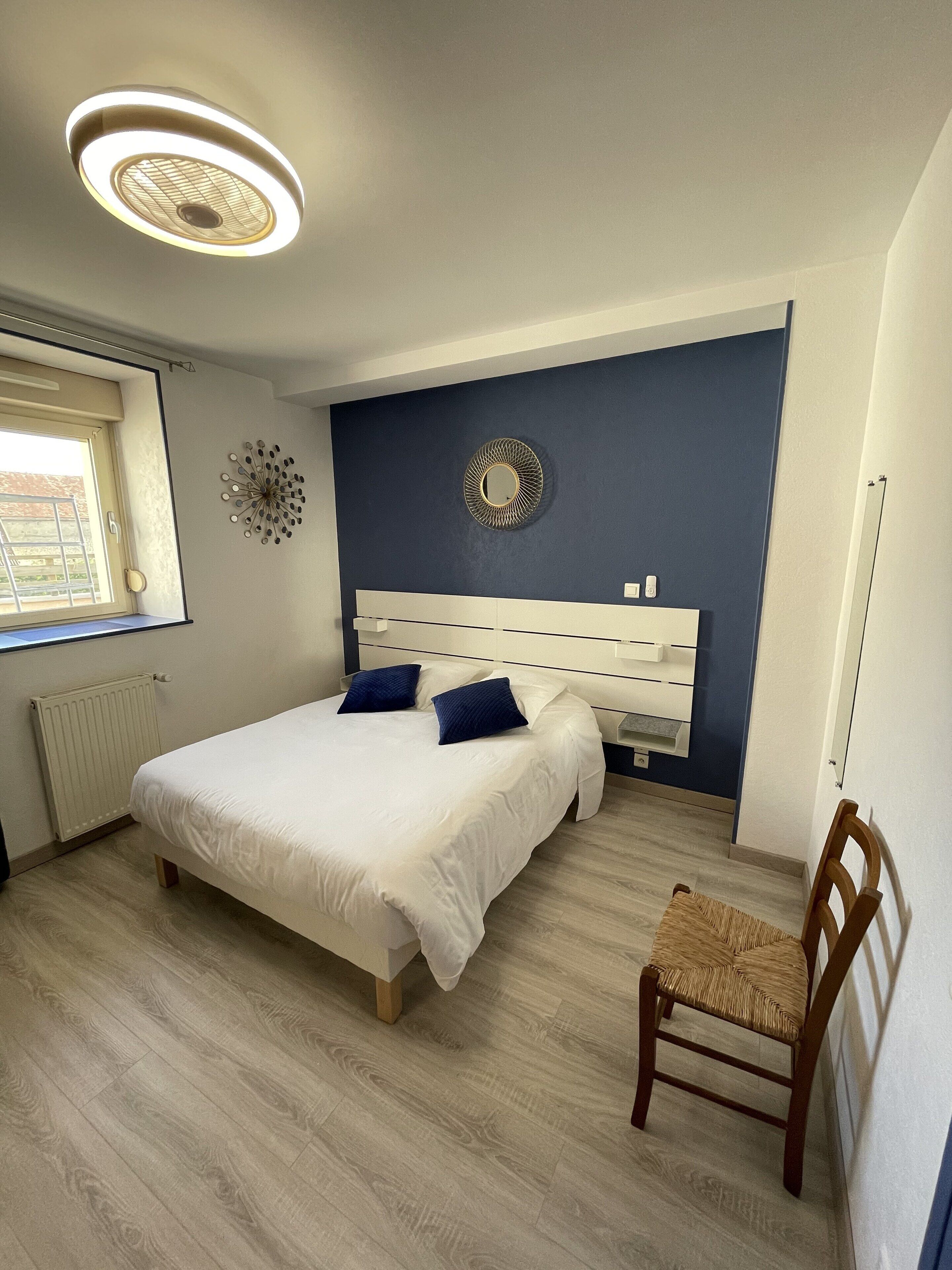Un dormitorio con cama, una silla, una ventana y un aplique de pared.
