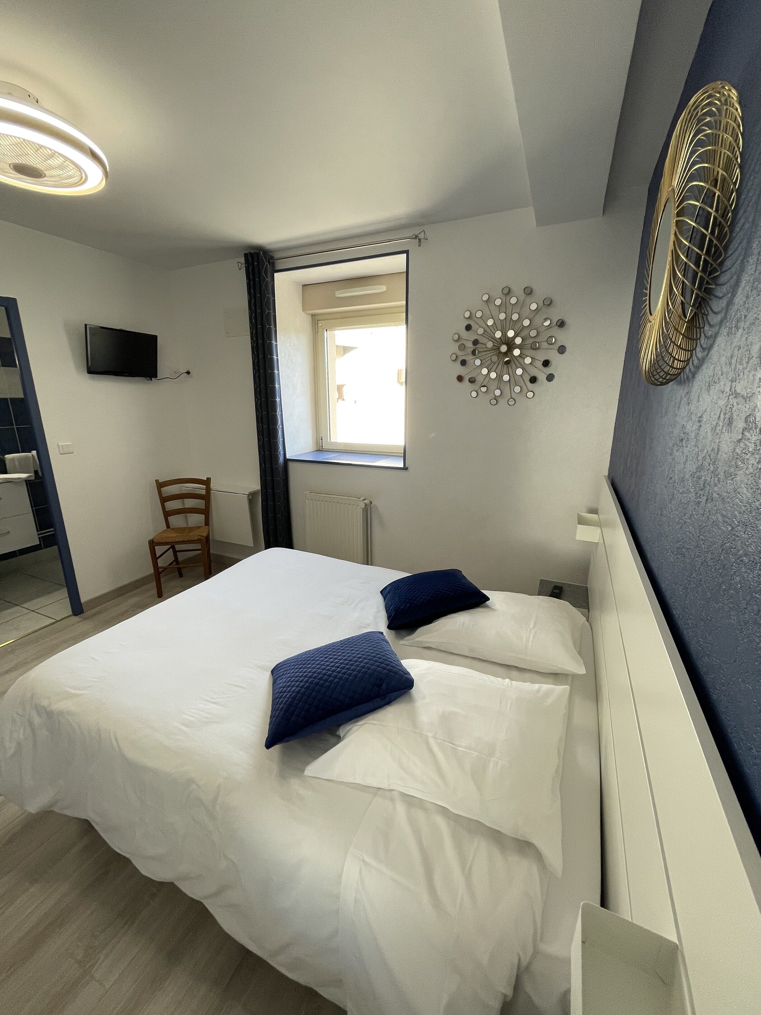 Une chambre à coucher comprenant un lit, deux oreillers, une chaise, un téléviseur, une fenêtre et un élément décoratif mural.