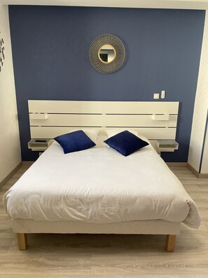 A neatly made bed with a white mattress and two blue pillows, a white headboard, and a wall-mounted mirror with a gold frame. - Hôtel Macarena (Pouillenay)