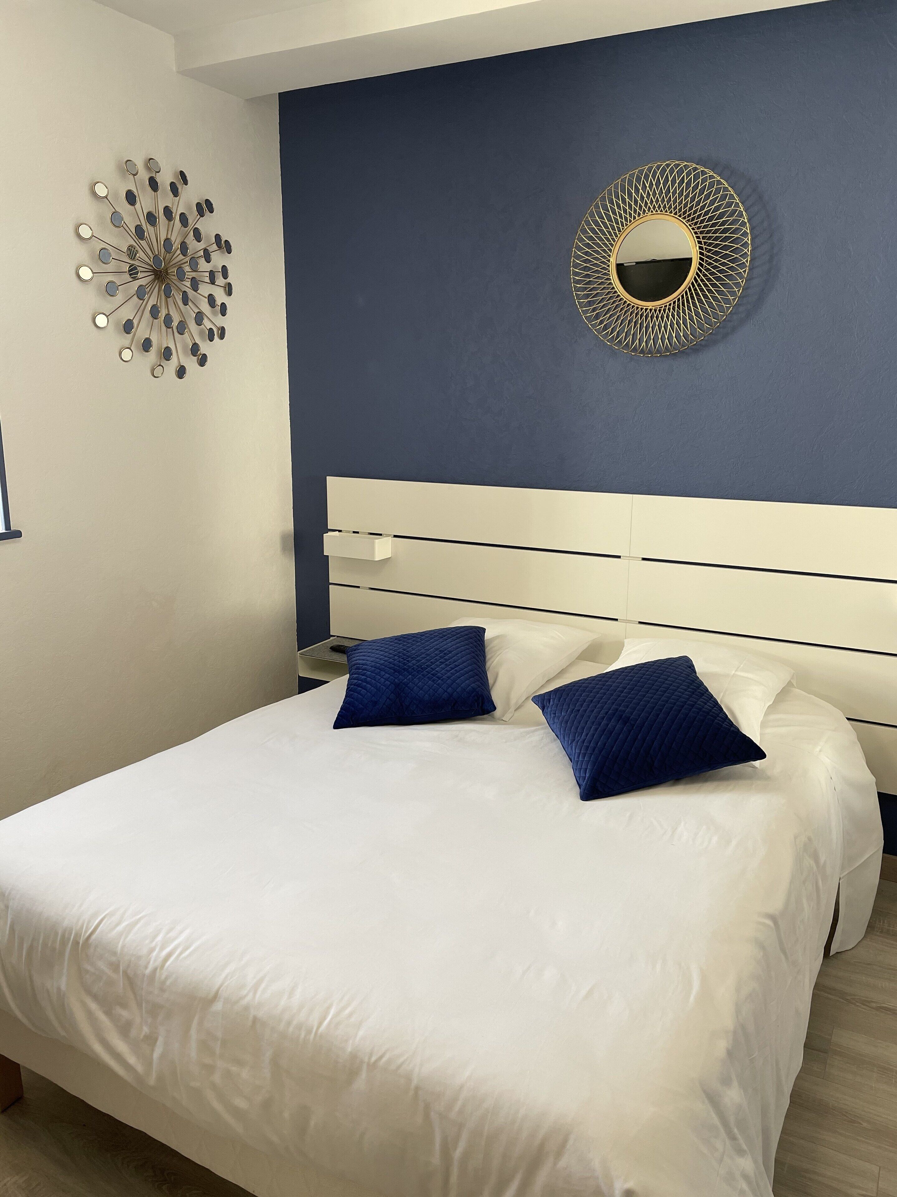 Ein Schlafzimmer mit einem weißen Bett, zwei blauen Kissen, einem weißen Kopfteil, einem runden Wandspiegel und einem dekorativen Wandbild.