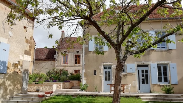 Una casa de dos pisos con jardín, un árbol y un pequeño espacio con muebles para disfrutar al aire libre.
