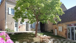 Un patio con un árbol, una mesa de madera y una silla. Se observa un edificio con una zona de recepción.