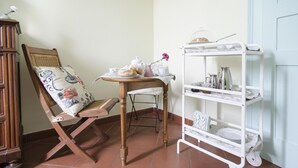 Une chaise en bois avec un coussin à motifs floraux, une petite table ronde recouverte d’une nappe blanche et une étagère métallique blanche contenant divers ustensiles de cuisine.