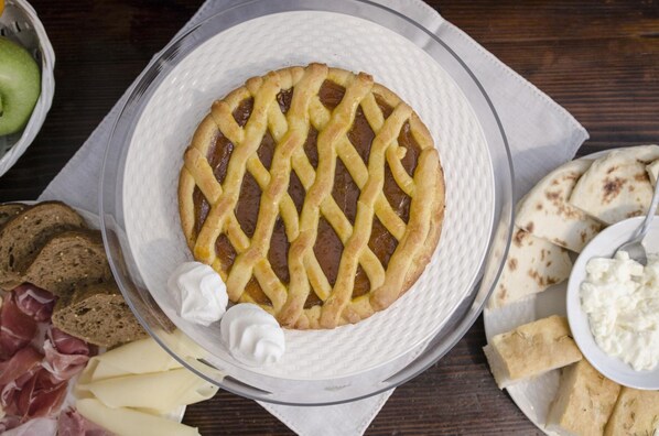 A pie with lattice crust on a plate, accompanied by various cheeses, bread, and a bowl of dip.