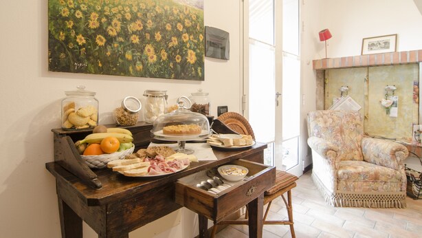 Une table à manger avec de la nourriture et des boissons, une chaise en osier et un fauteuil à motifs floraux dans une pièce où un tableau est accroché au mur.