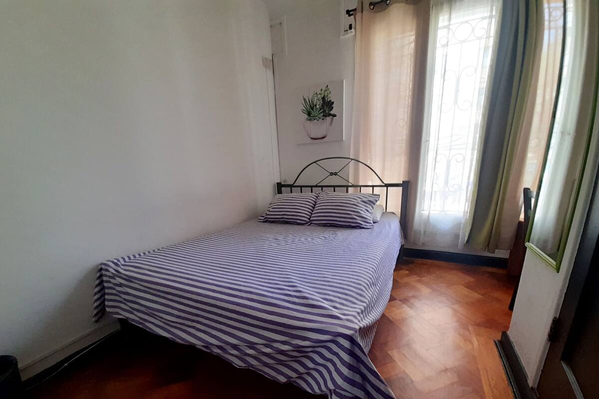 A single bed with a striped bedspread in a room with wooden flooring, a potted plant on the wall, and a window with curtains.