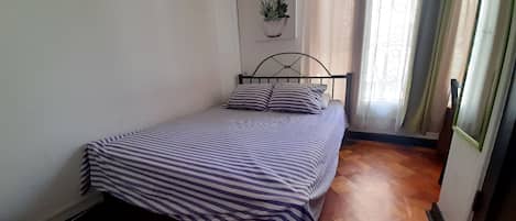 Un lit simple avec une couvre-lit à rayures, dans une pièce au plancher en bois, une plante en pot accrochée au mur et une fenêtre avec des rideaux.