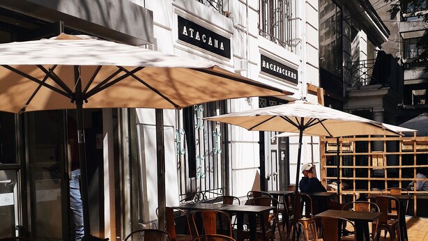 An outdoor cafe with tables and chairs, umbrellas, and a building with signs reading "ATACAMA" and "BACKPACKERS". - Atacama Backpackers (Santiago)