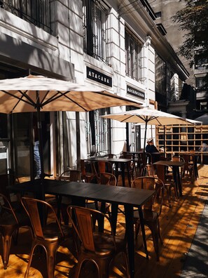 An outdoor cafe with tables and chairs, umbrellas, and a building with signs reading "ATACAMA" and "BACKPACKERS". - Atacama Backpackers (Santiago)