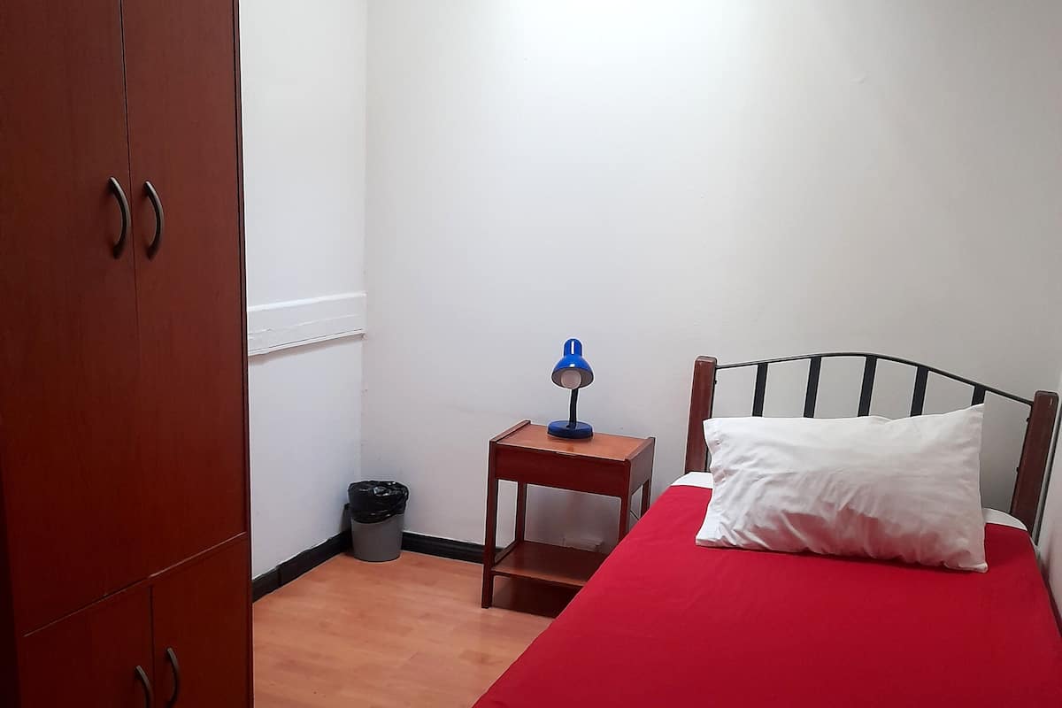 A bedroom with a bed, a wooden wardrobe, a small table with a lamp, and a trash bin.