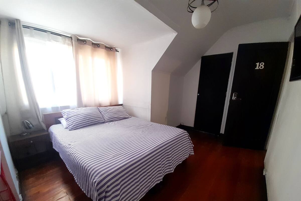 A bedroom with a bed, a nightstand, a window with curtains, a door, and a ceiling light.