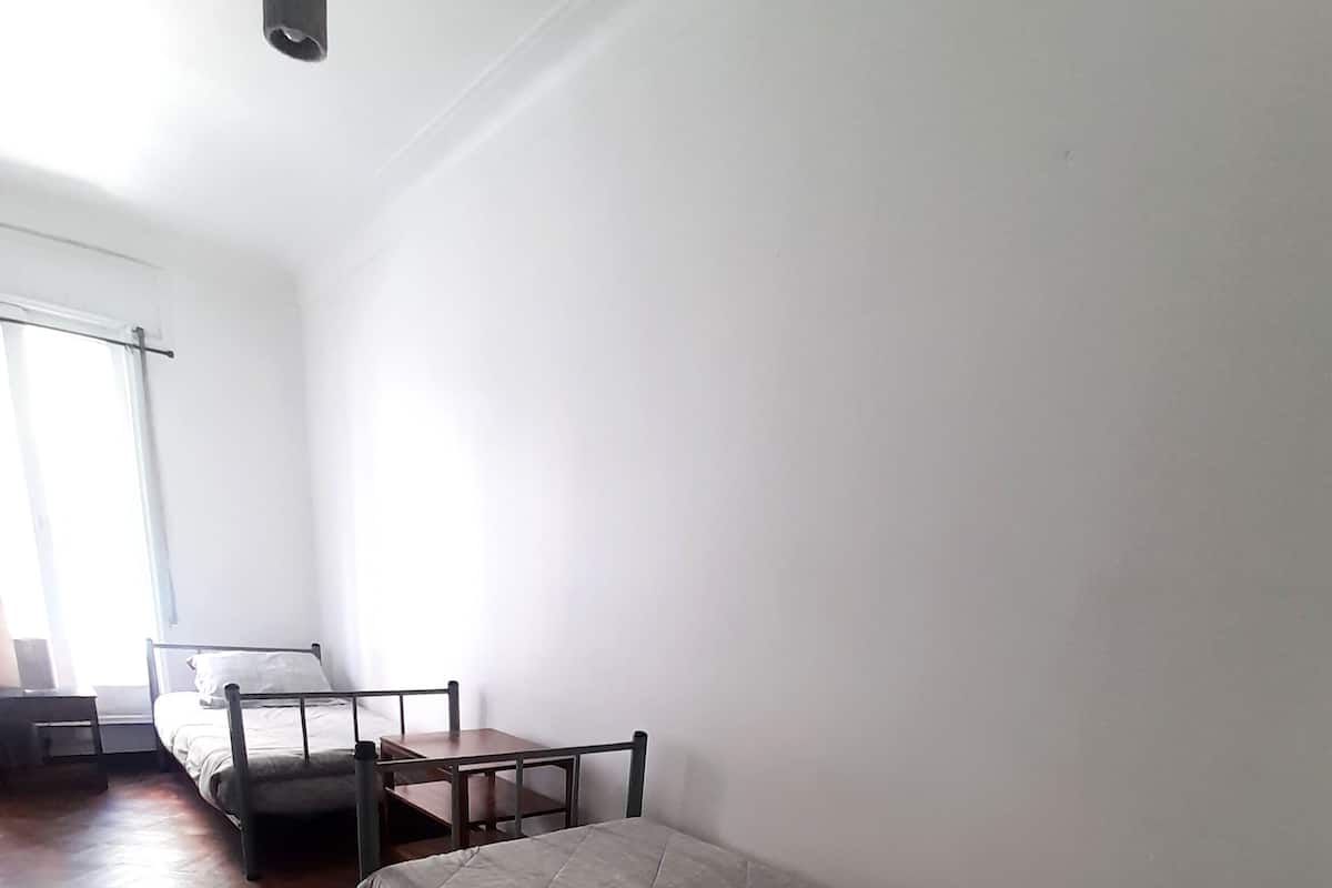 A room with a wooden floor, a single bed with a quilted mattress and pillows, a small wooden table, and a chair.