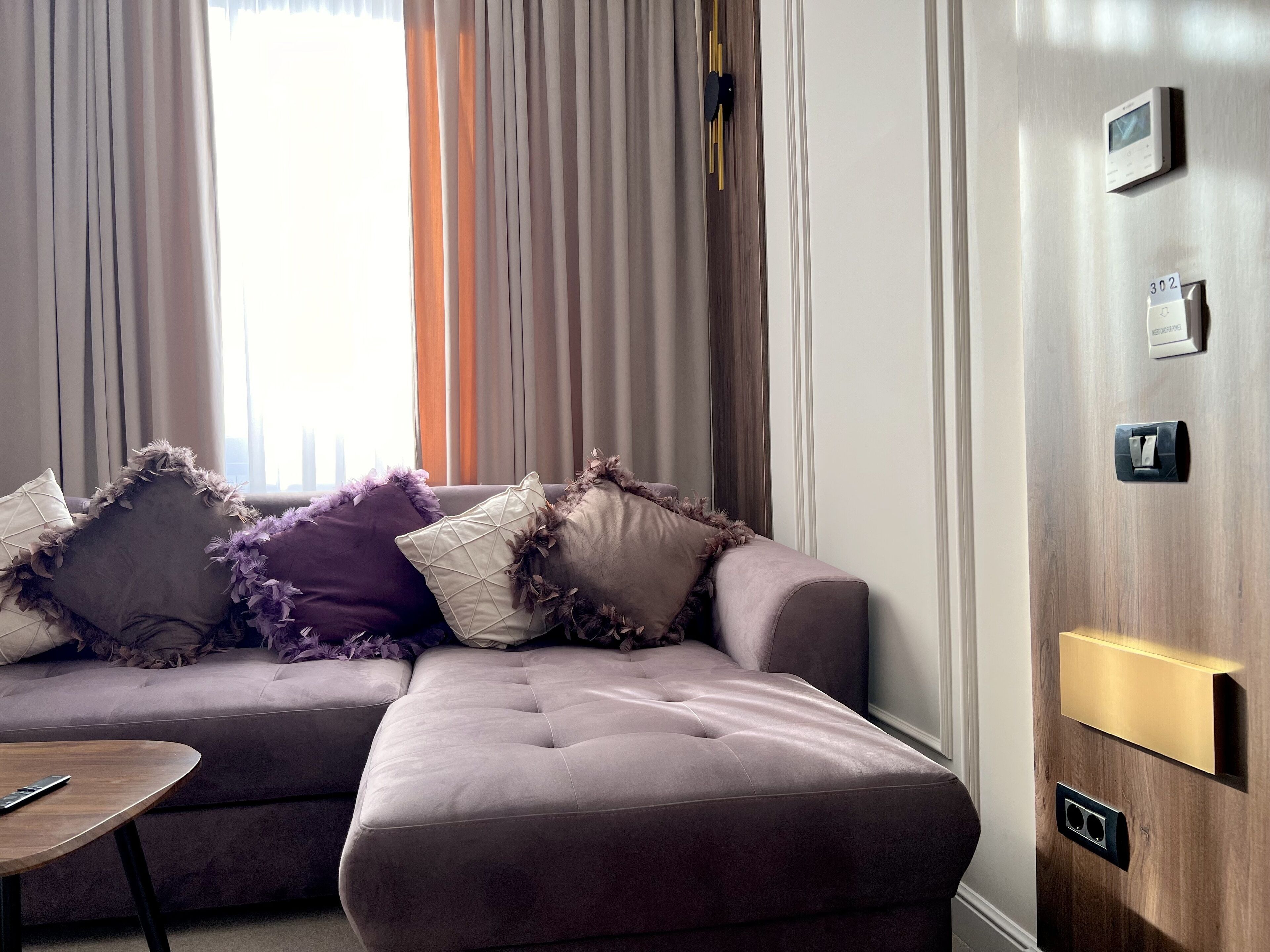 A modern living room with a sectional sofa, a small wooden table, and a wall-mounted thermostat.