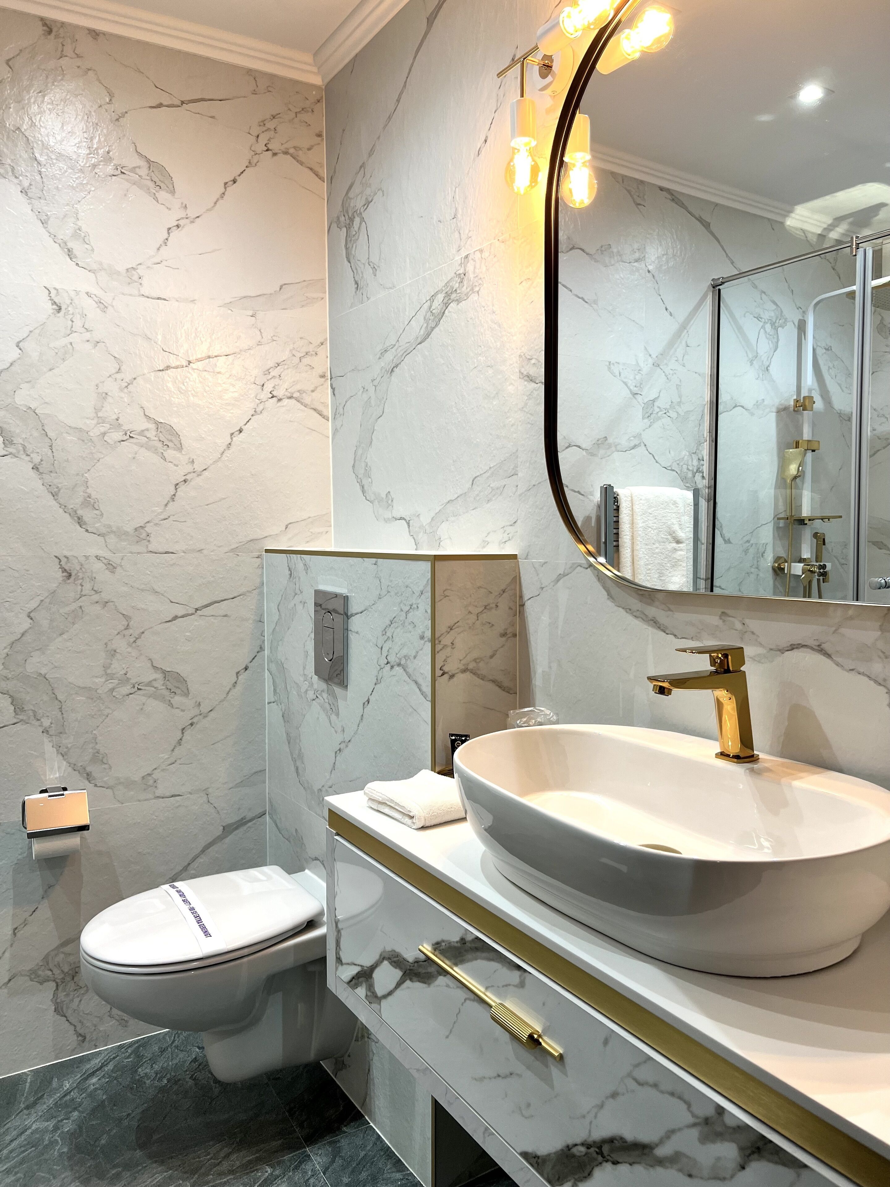 A modern bathroom with a marble wall, a large mirror, a white sink, and a toilet.