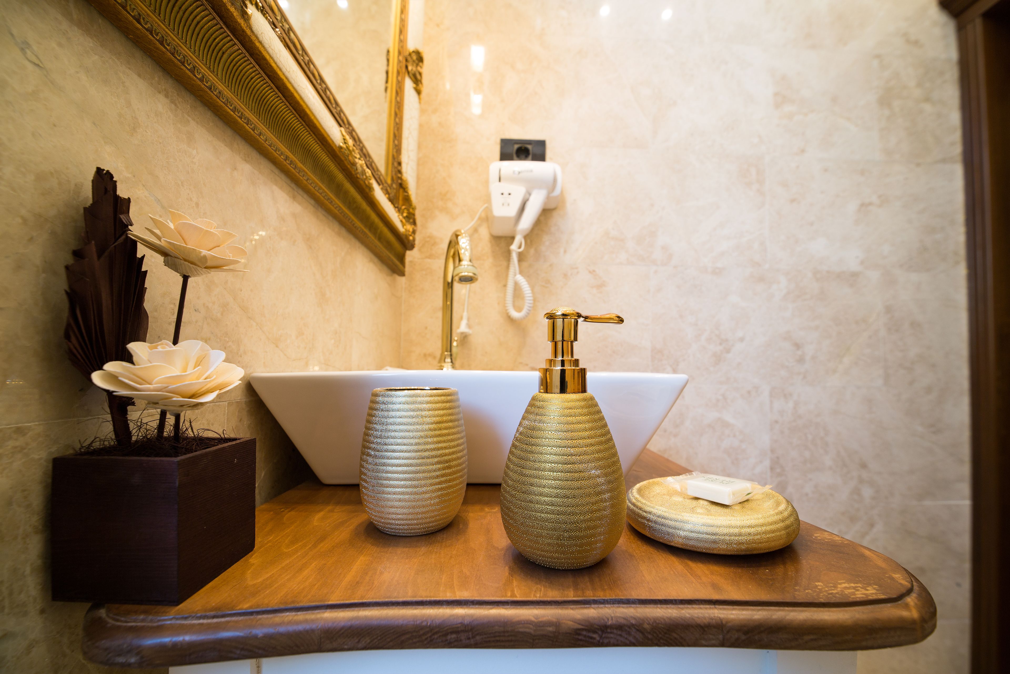 Un tocador de baño con lavamanos blanco, grifo dorado y elementos decorativos como un florero y un jabonera.