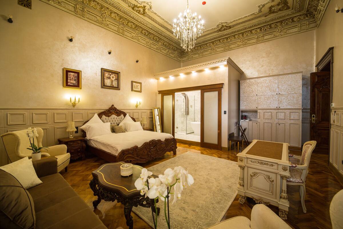 A spacious bedroom with a large bed, a sitting area with a sofa and armchair, a small table, and a decorative mirror.