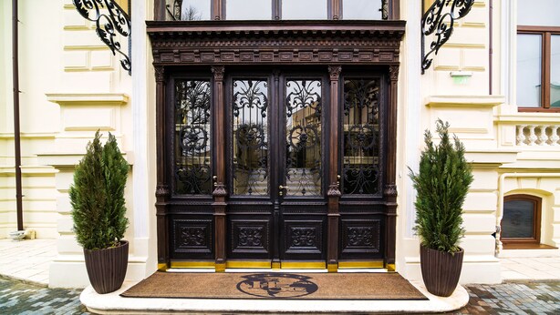 A grand entrance with a dark wooden door, two potted plants, and a doormat. - Hotel Boutique 5 Continents (Craiova)