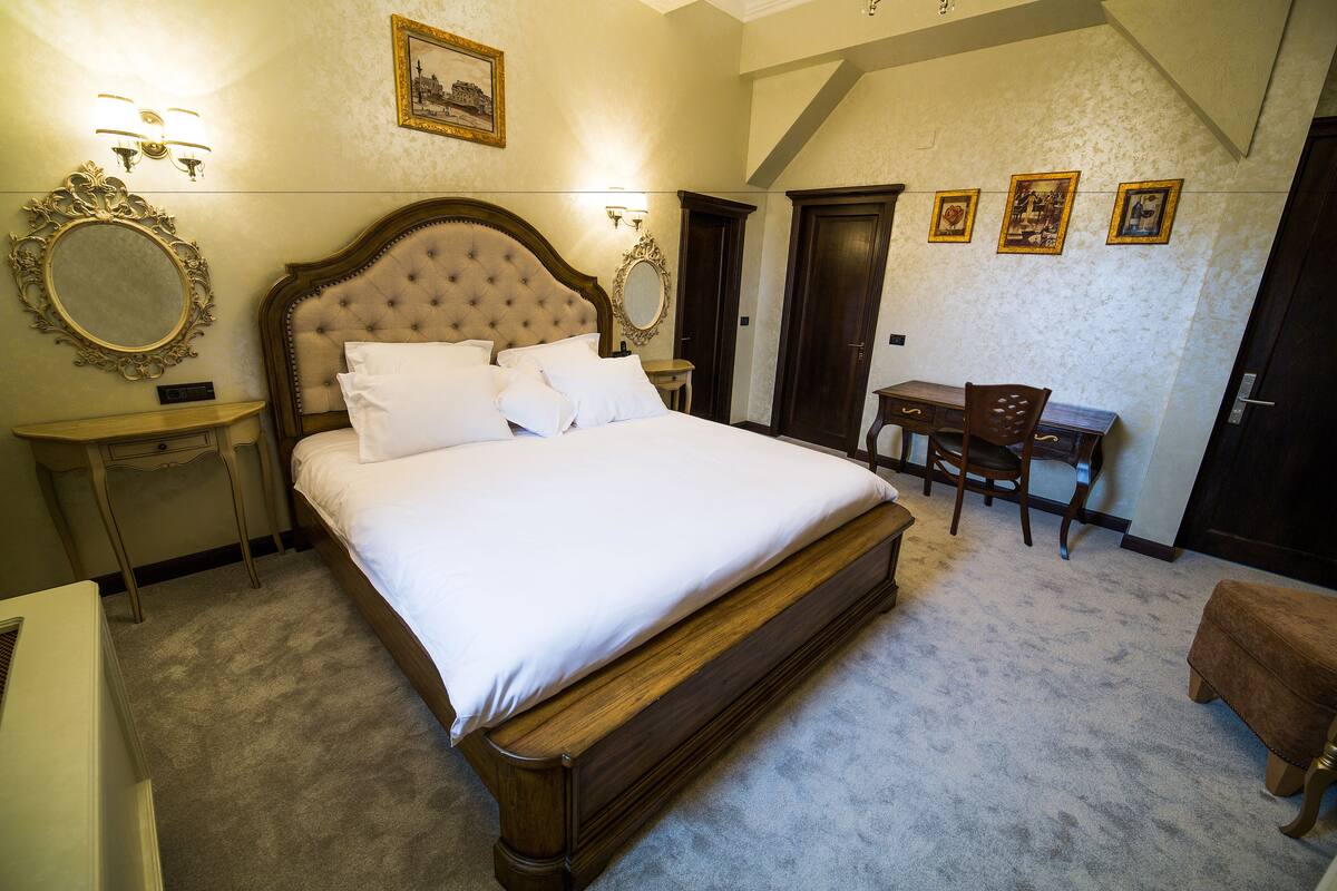 A bedroom with a large bed, a desk, and a chair. There are two bedside tables, a mirror, and framed pictures on the wall.