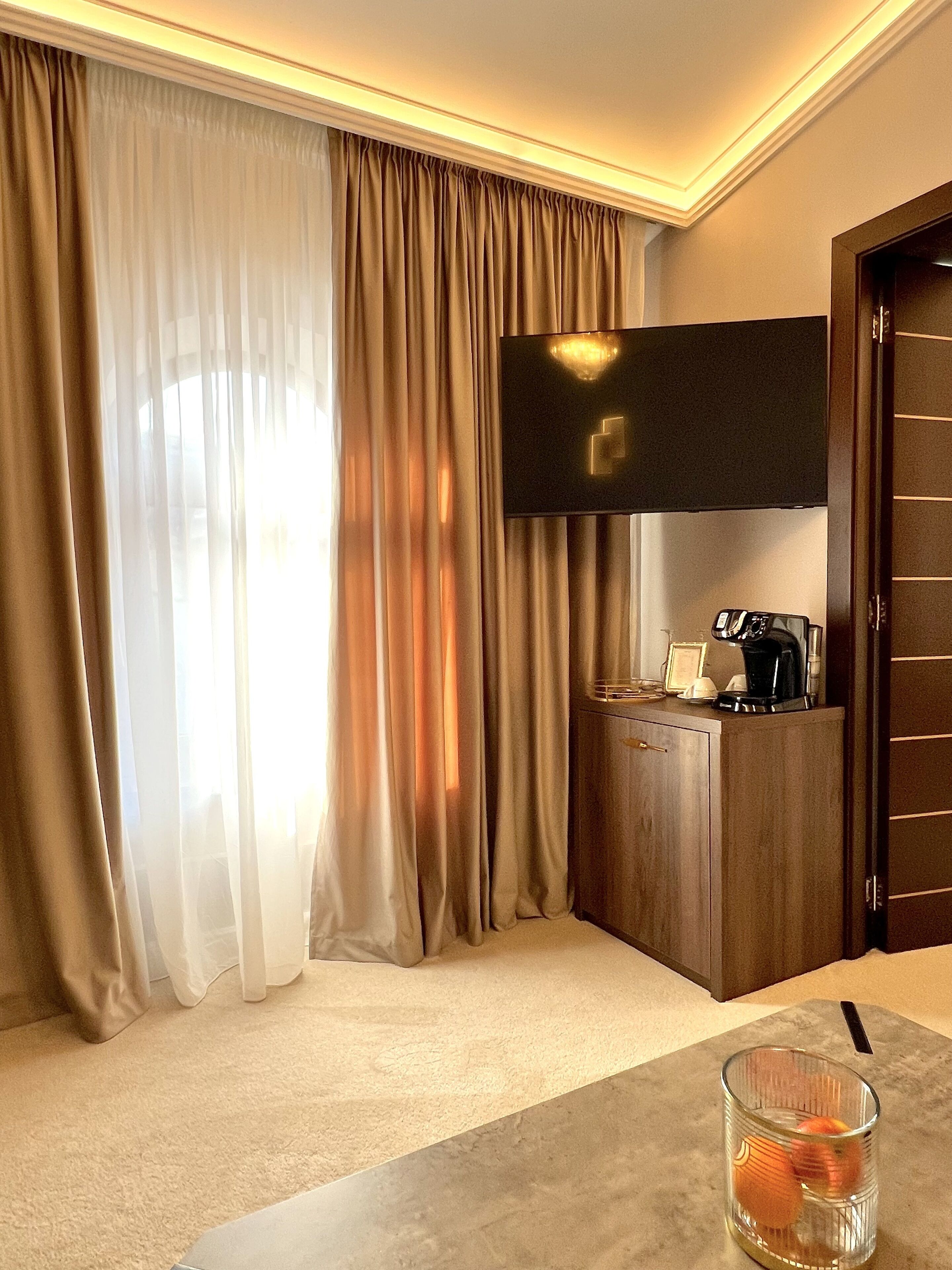 Ein Hotelzimmer mit großem Fenster, einem flachbildfernseher an der Wand, einem Holzschrank und einem Glastisch mit einer Tasse.