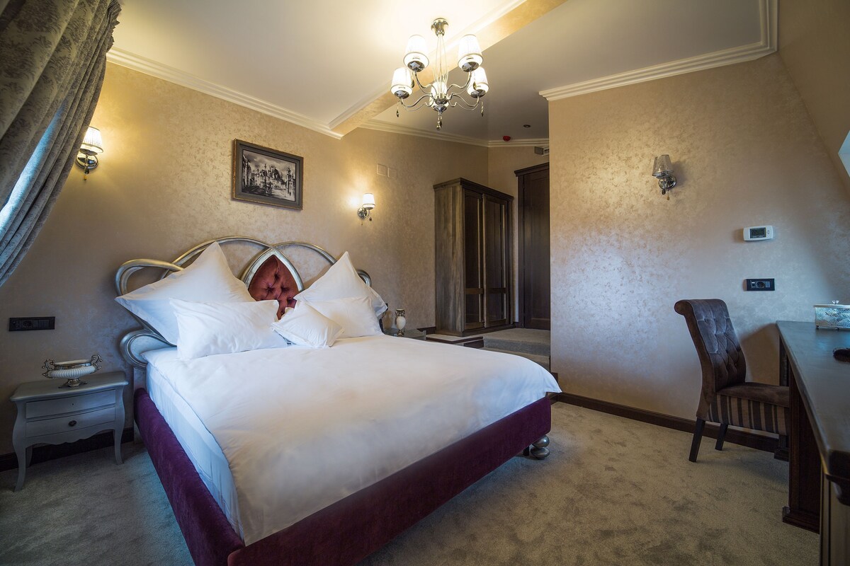 Ein Hotelzimmer mit einem großen Bett, einem Schreibtisch, einem Sessel, einer Kristallleuchtre und einer Tür zu einem weiteren Raum.
