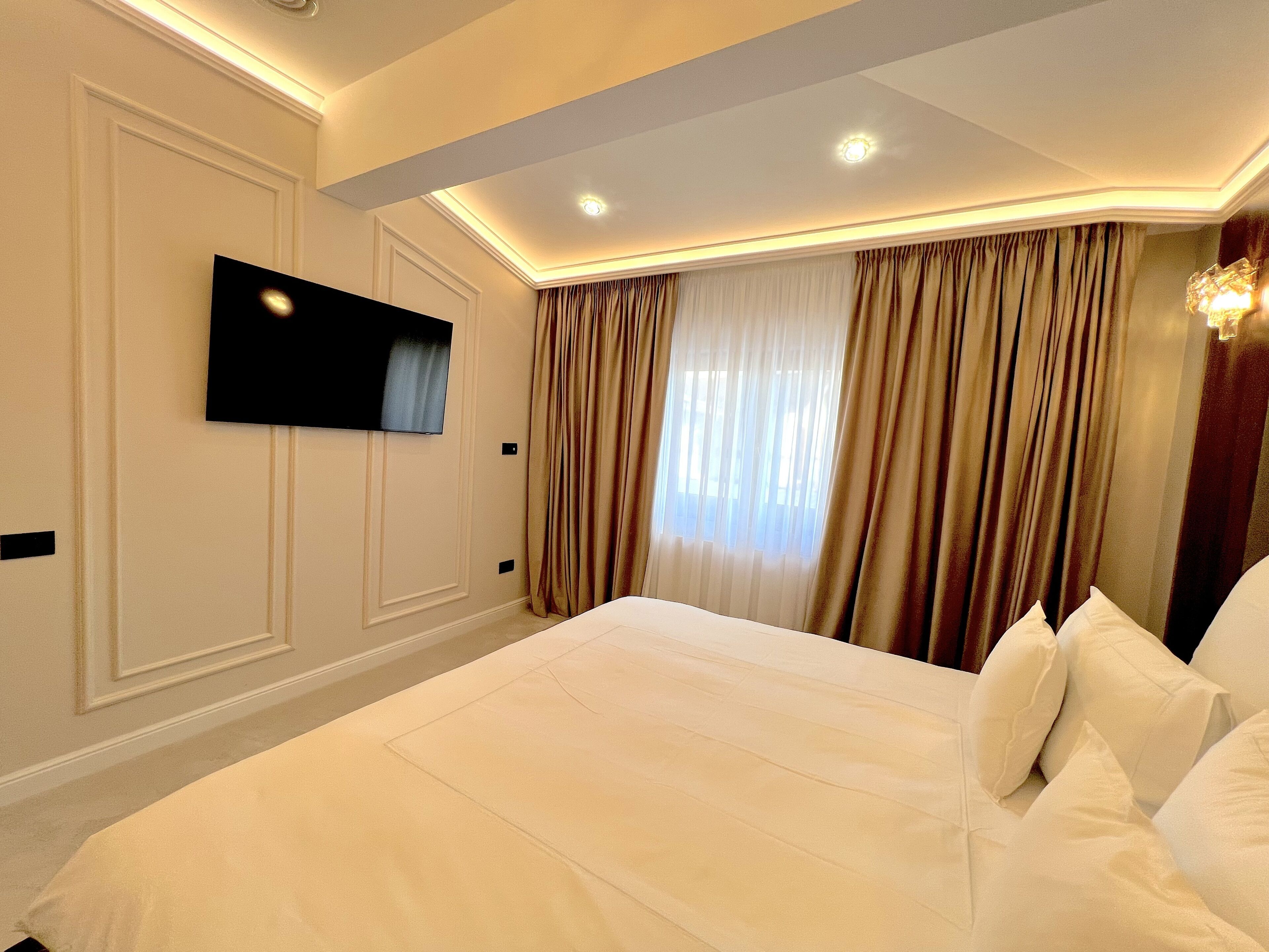 A hotel room with a large bed, a flat-screen TV mounted on the wall, and a window with curtains.