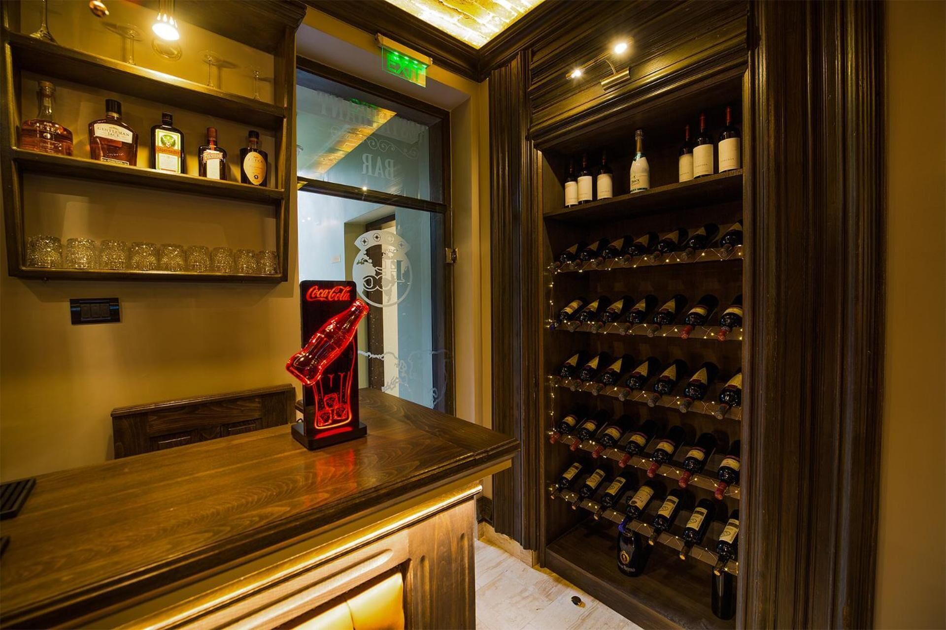 Zona de bar con estantes llenos de botellas de licor, un letrero de neón de Coca-Cola y una vinoteca con varias botellas.