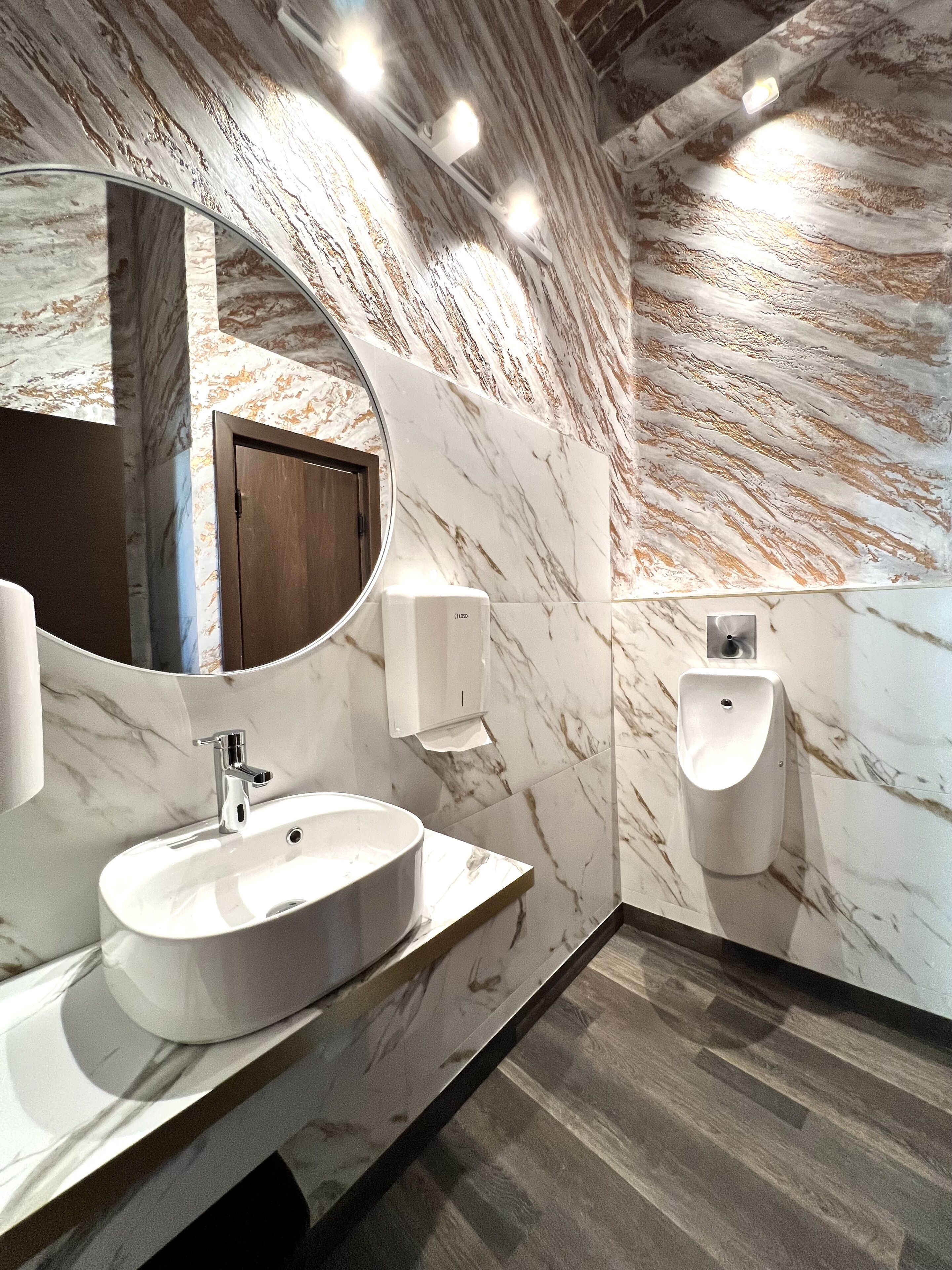Un baño moderno con tocador de mármol, espejo redondo, puerta de madera y orinal.