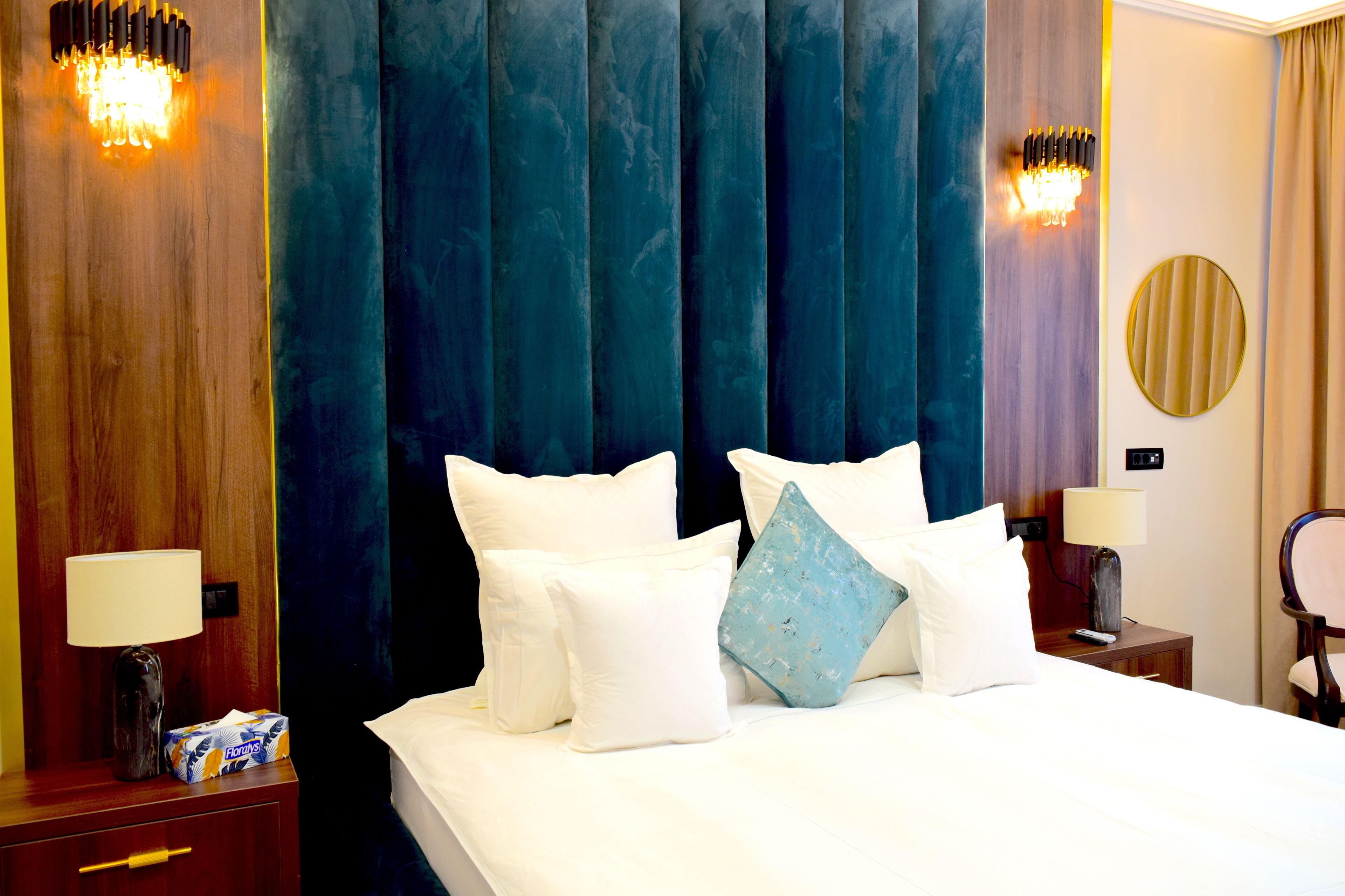 A hotel room with a large bed, white bedding, a wooden nightstand with a lamp, a tissue box, and a round mirror on the wall.