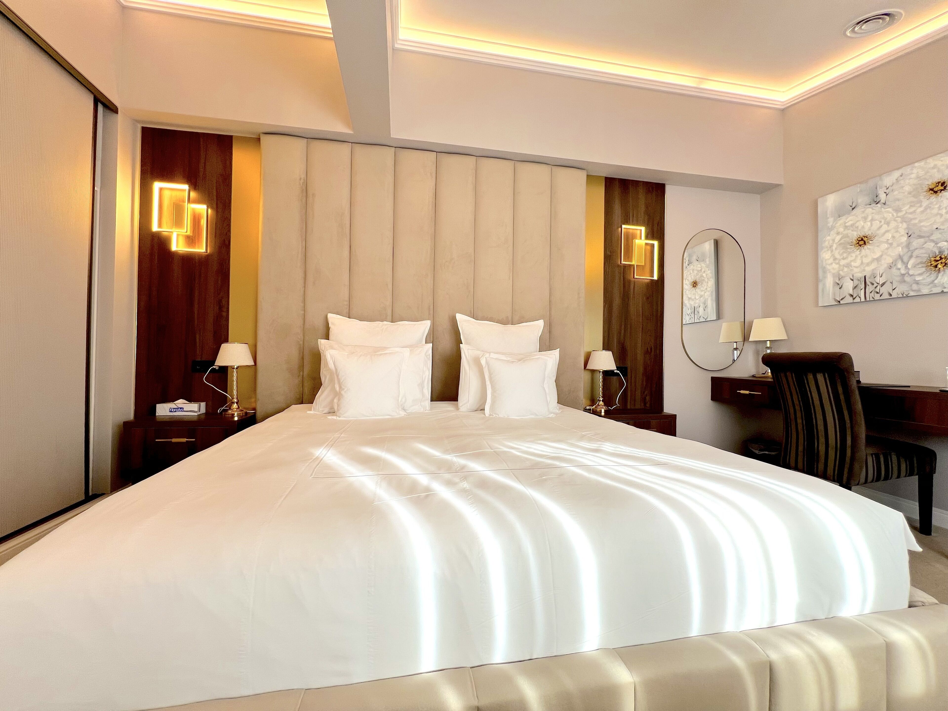 A hotel room with a large bed, two bedside tables with lamps, a wooden headboard, a desk with a chair, and a large painting on the wall.