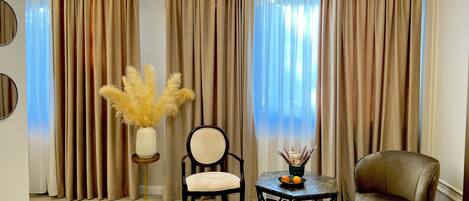 A room with beige curtains, a round mirror, a vase with dried plants, a chair, a small table with fruits, and a plush armchair.