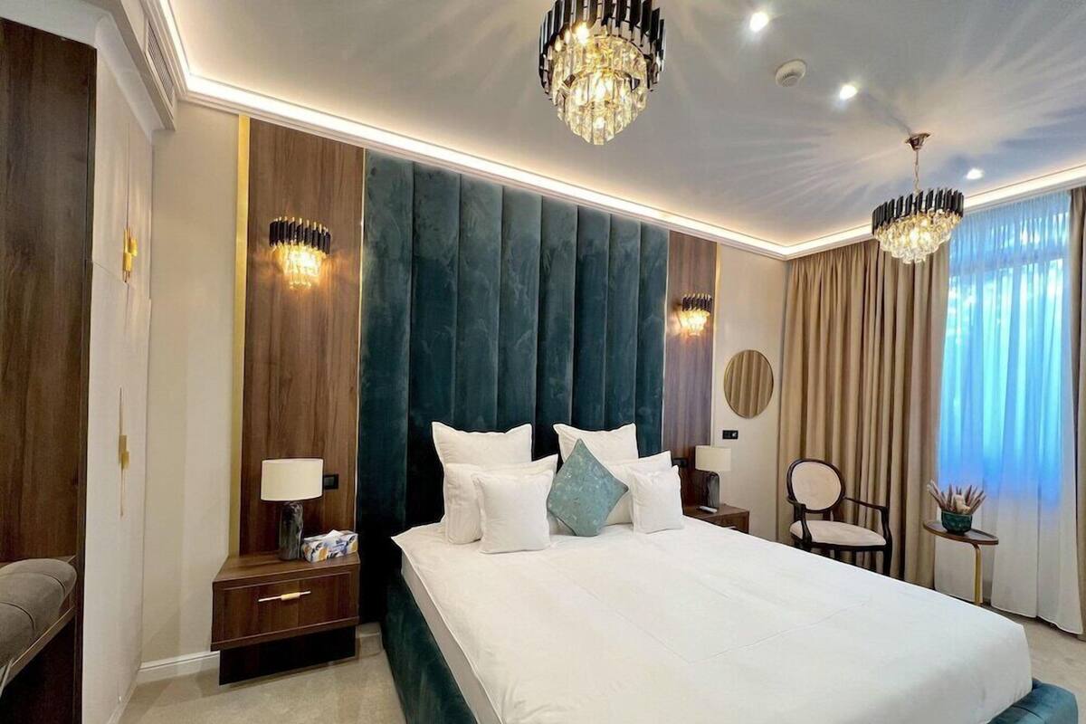 A modern hotel room with a large bed, two bedside tables, a chair, a round mirror, and a chandelier.