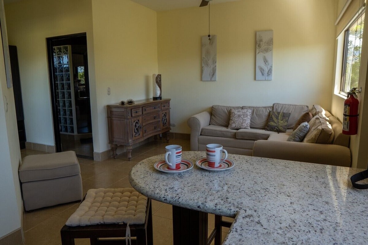 Una sala acogedora con una mesa redonda de mármol, dos sillones con cojines, un sofá color beige y un mueble de madera.