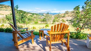 Una terraza de madera con dos sillas Adirondack y una mesita, con vistas a un paisaje exuberante con colinas y árboles.