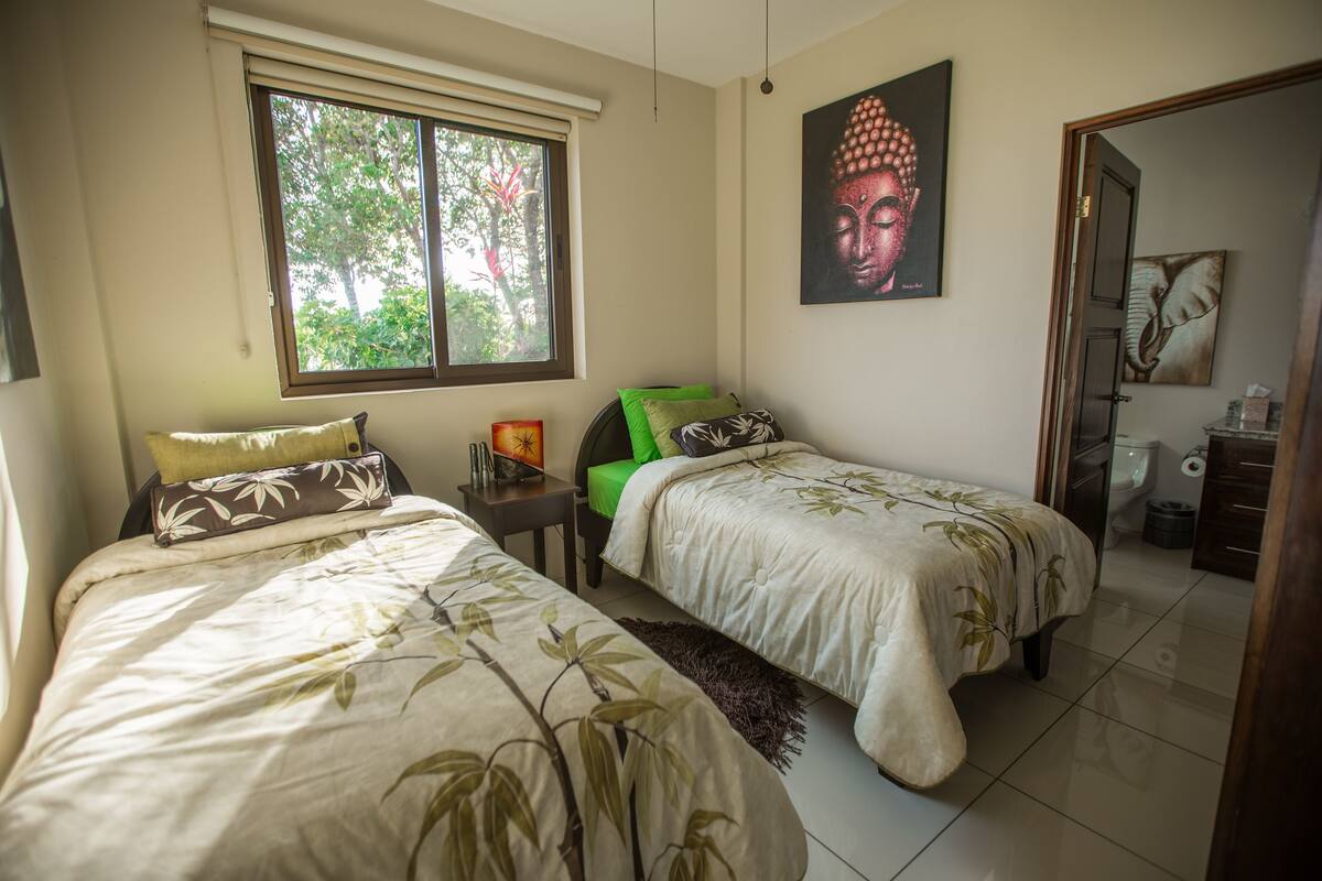 Un dormitorio con dos camas, una ventana, un cuadro de Buda y un baño visible a través de una puerta abierta.