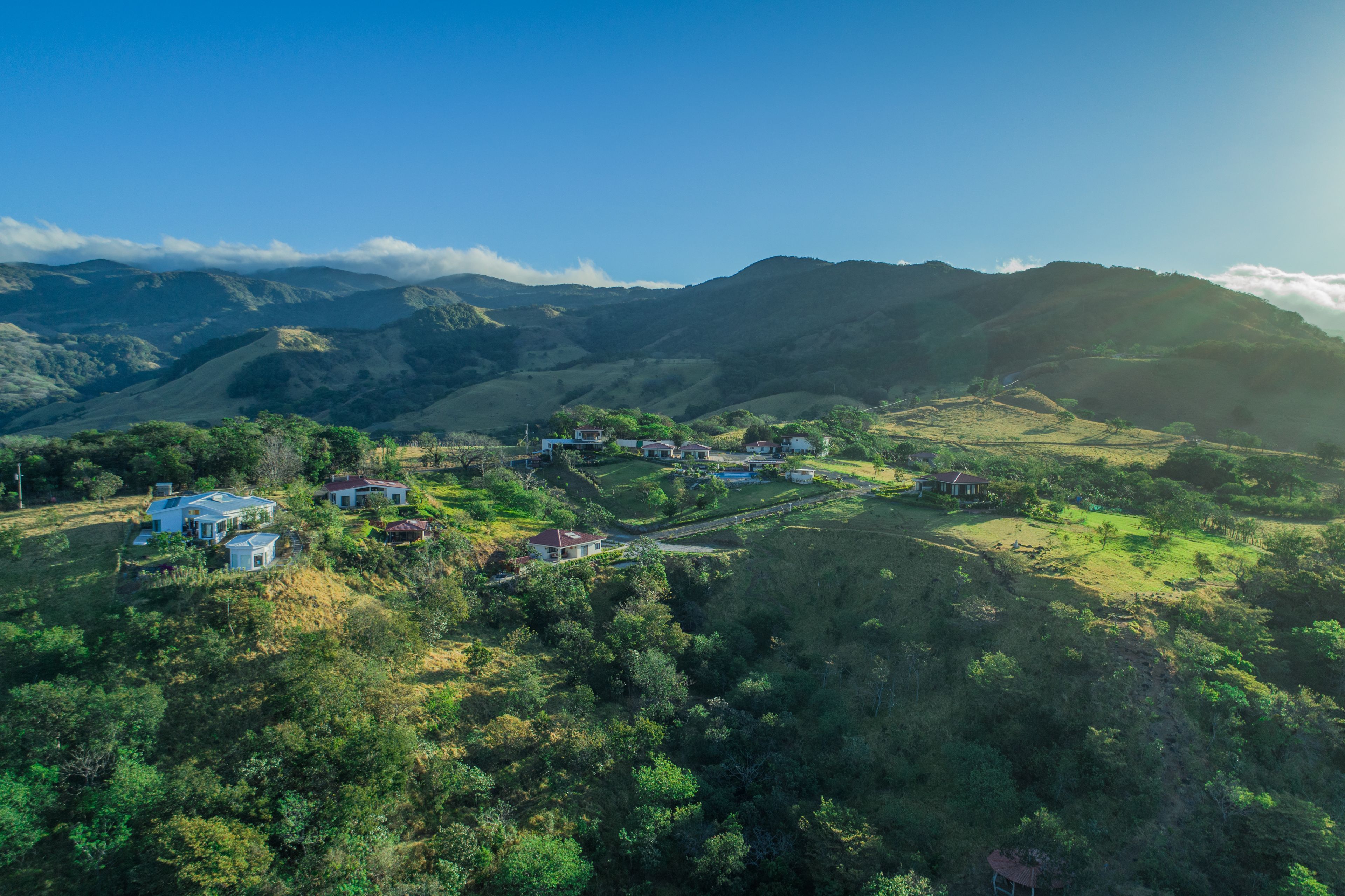 An aerial view of a rural area with several houses surrounded by lush greenery and hills.