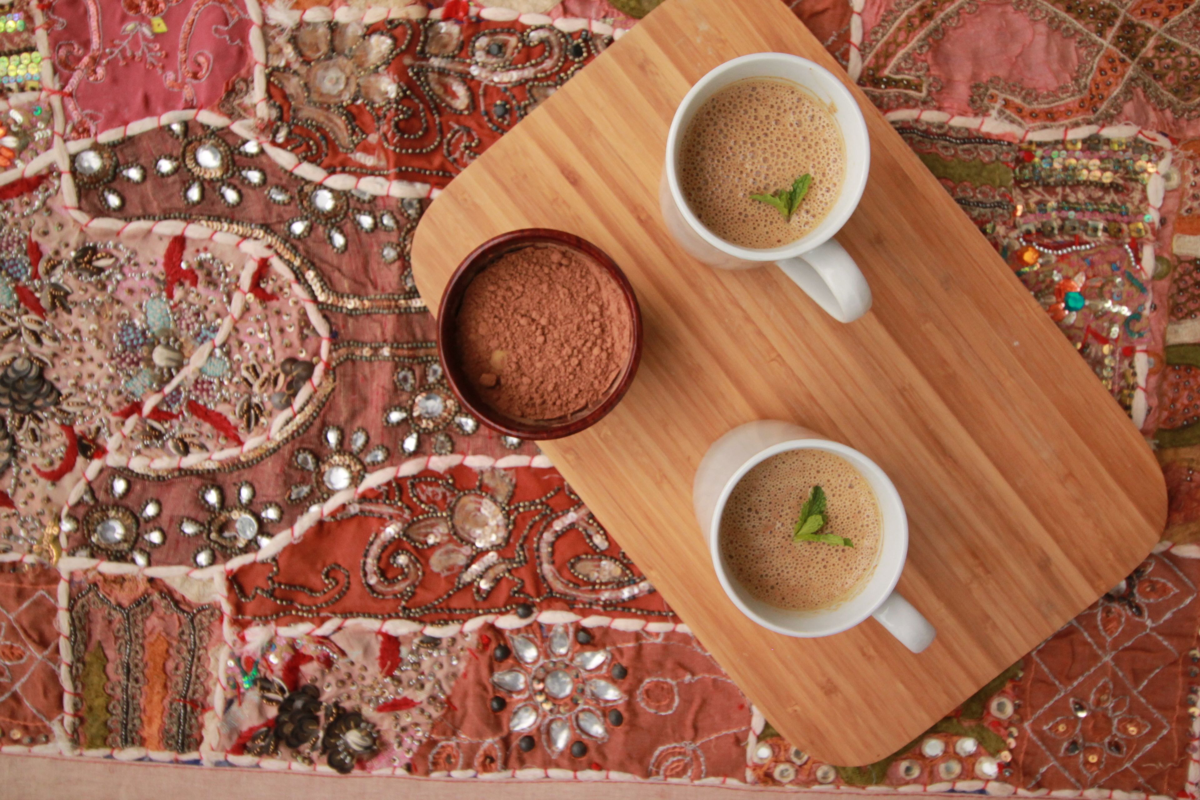 Two white cups of coffee on a wooden tray, placed on a patterned fabric.