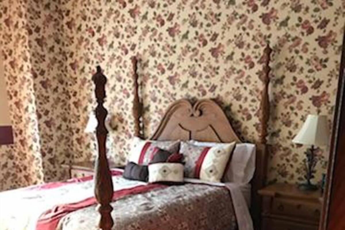 A bedroom with a four-poster bed, floral wallpaper, and a nightstand with a lamp.