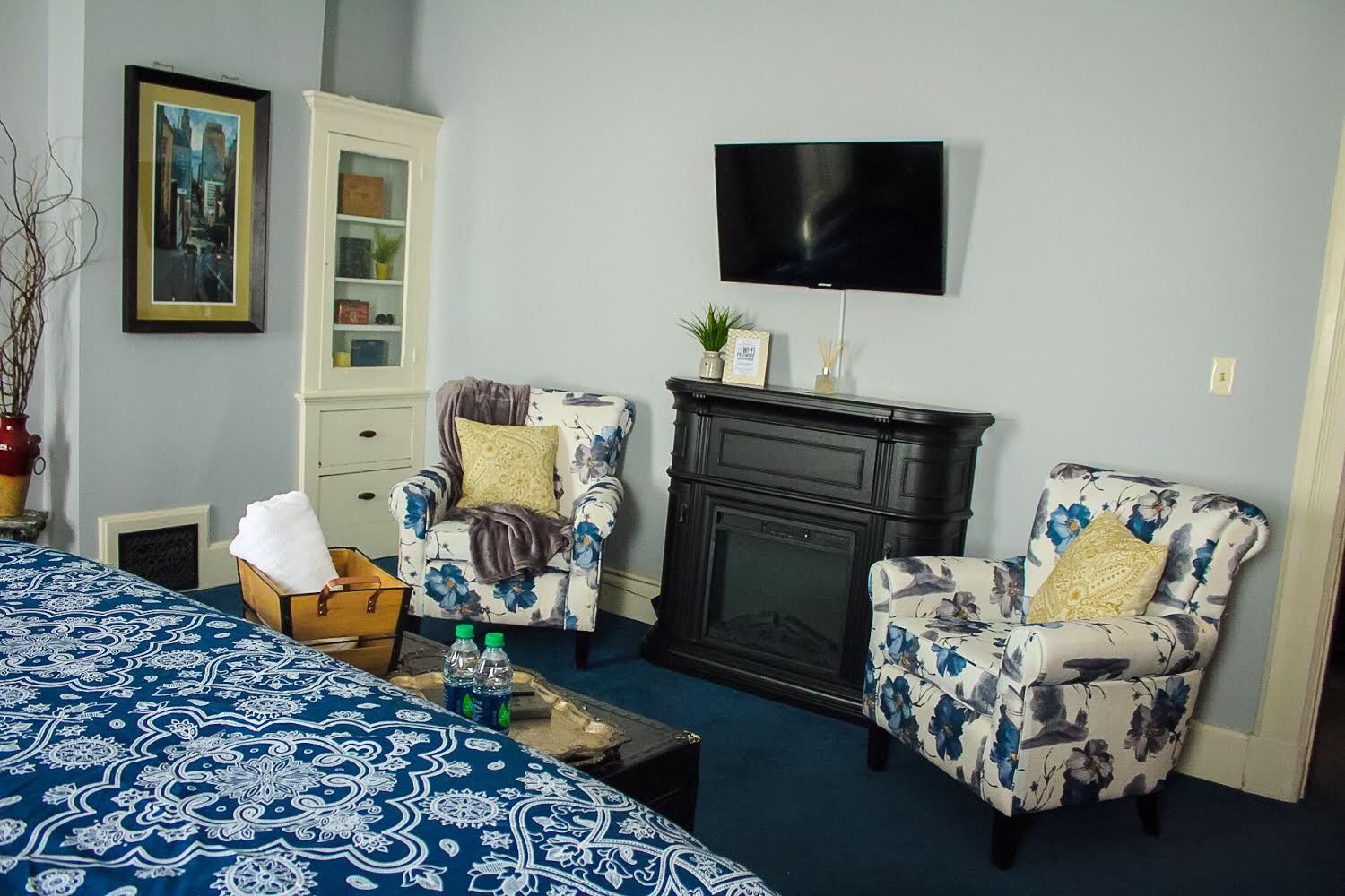 A room with a bed, two floral armchairs, a television, a fireplace, and a cabinet with glass doors.