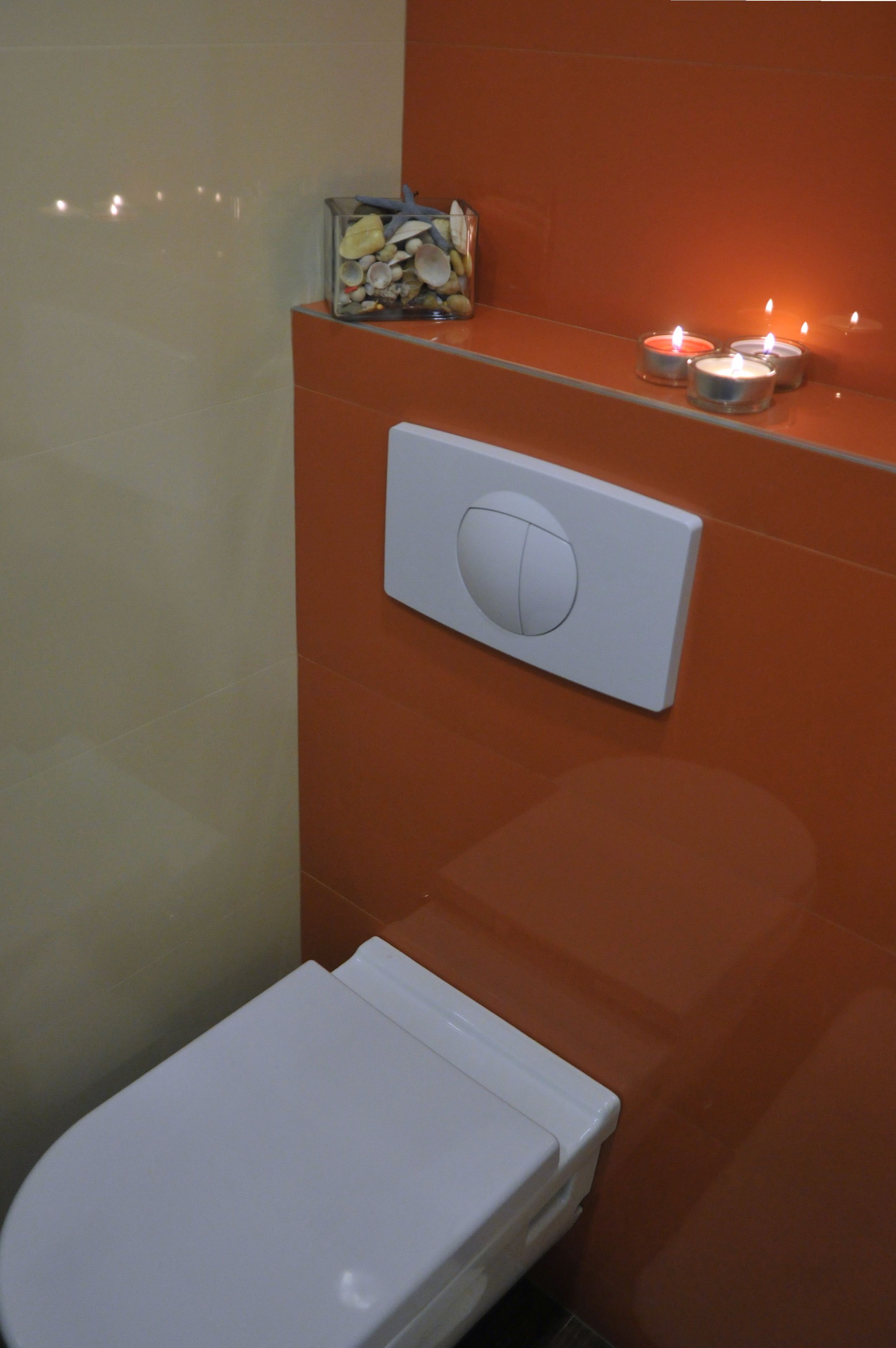 Ein Badezimmer mit einer weißen Toilette, einer orangefarbenen Wand, einem Lichtschalter und einer kleinen Schale mit Meeresschnecken.