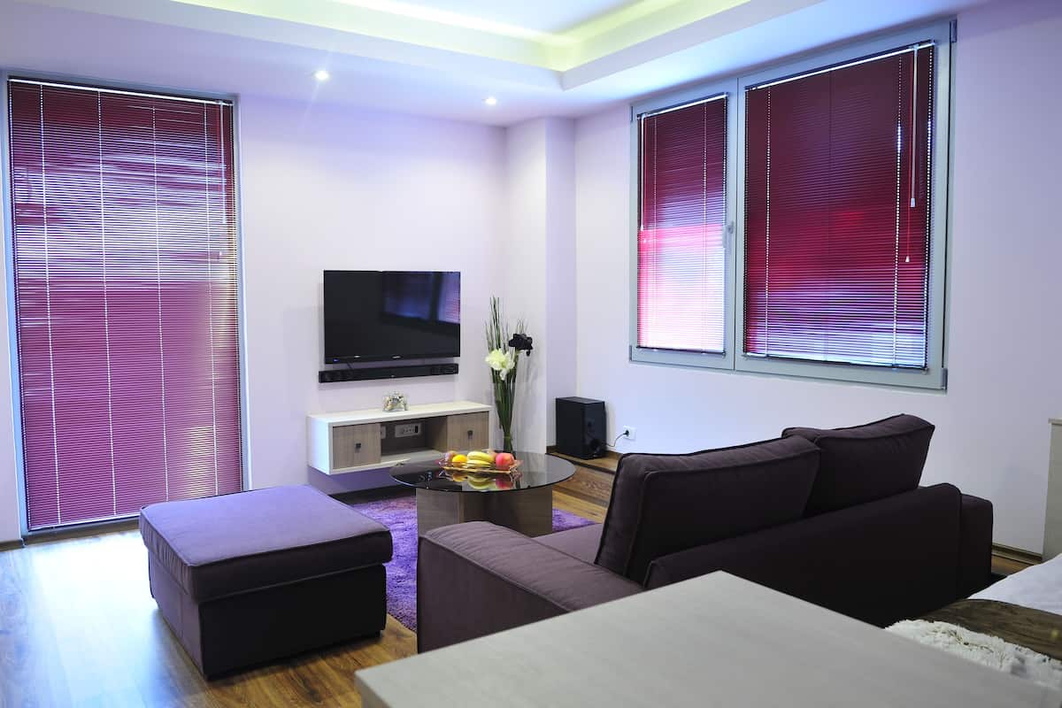 A modern living room with a flat-screen TV, a sofa, a coffee table with fruit, and large windows with blinds.