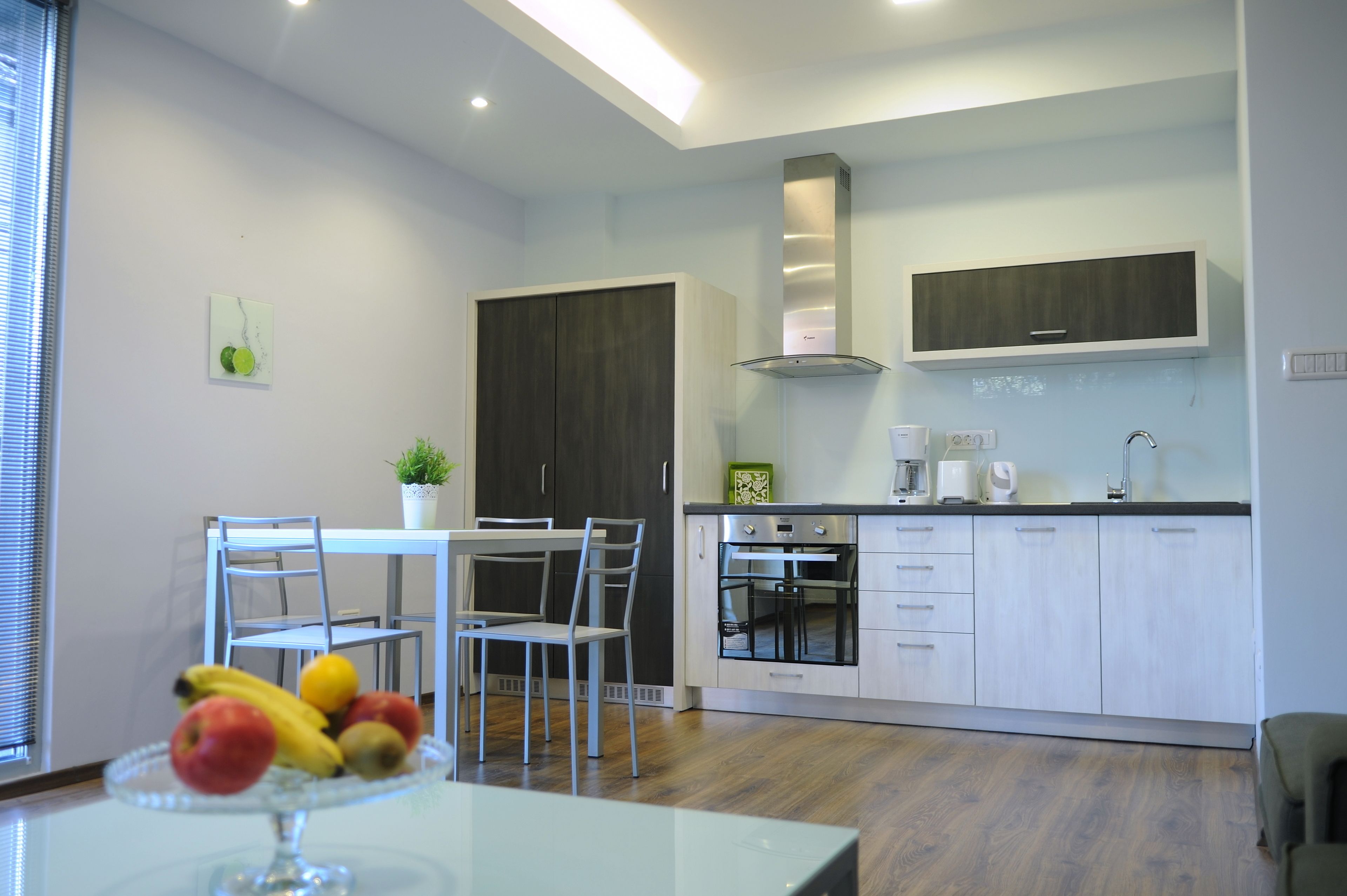 A modern kitchen with white cabinets, a built-in oven, and a stainless steel range hood. There is a dining area with a table and chairs, and a fruit bowl on the table.