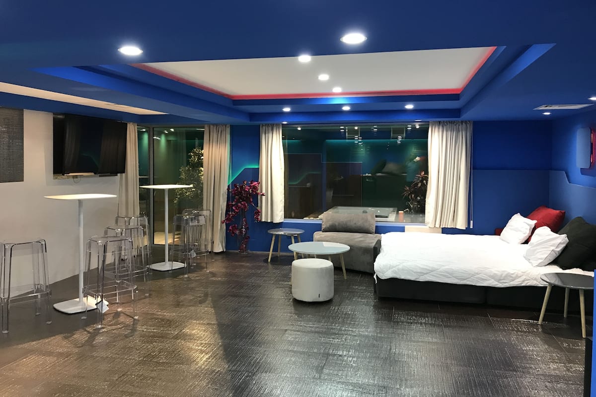 A modern hotel room with a large bed, a sofa, a coffee table, and a TV. There is a balcony with a view of the city.