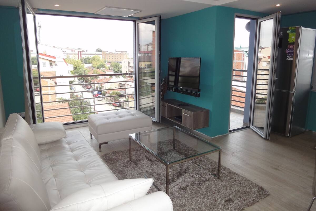 A modern living room with a white sofa, a glass coffee table, and a TV mounted on the wall.
