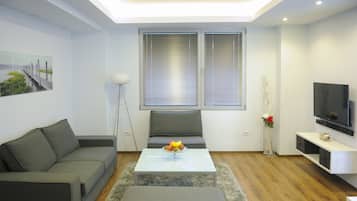 Una sala de estar moderna con un sofá gris, una mesa de centro de vidrio con fruta, un sillón gris, un televisor sobre un mueble blanco y un cuadro en la pared.