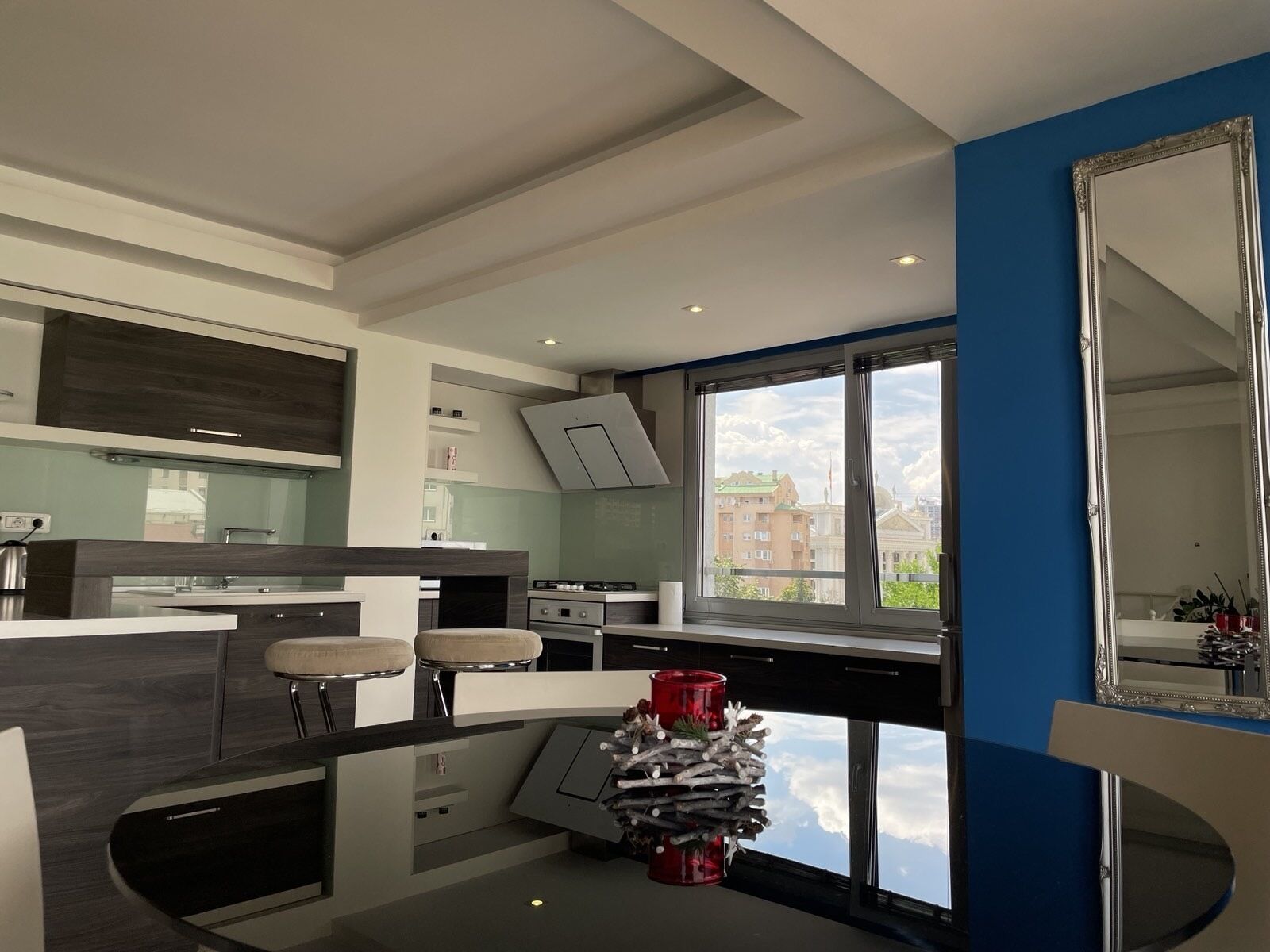 A modern kitchen with a large window, a dining area with bar stools, and a decorative plant centerpiece.