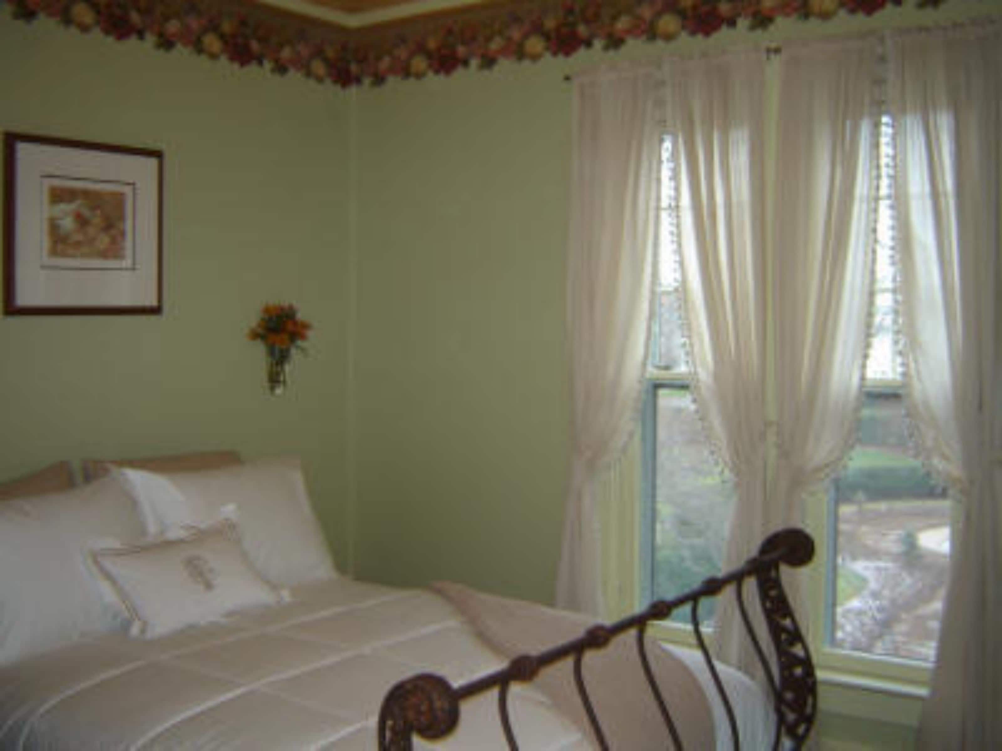 Un dormitorio con una cama, una ventana con cortinas, un cuadro enmarcado en la pared y un florero con flores.
