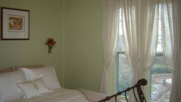 Un dormitorio con una cama, una ventana con cortinas, un cuadro enmarcado en la pared y un florero con flores.