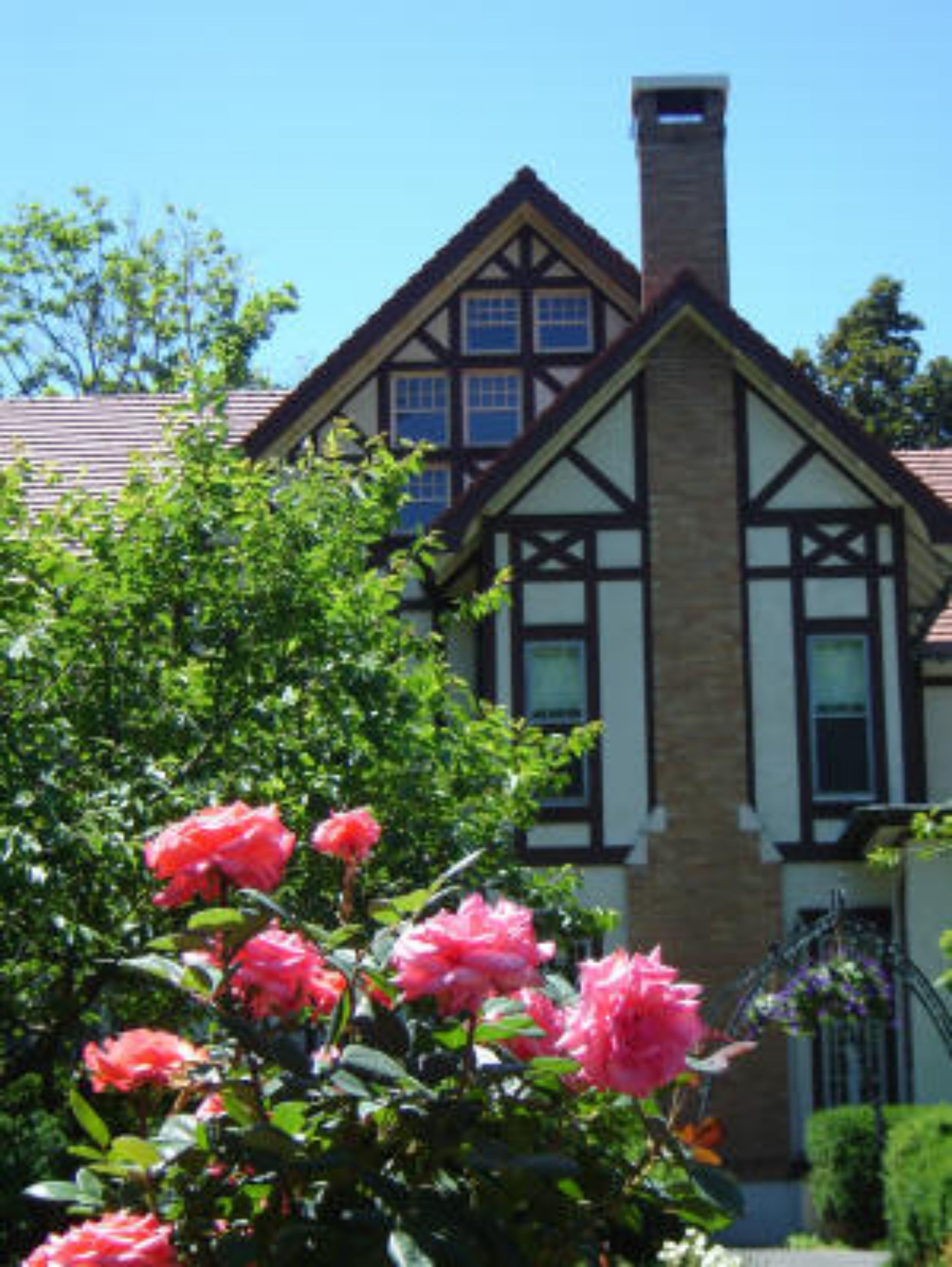 A Tudor-style house with a prominent chimney, large windows, and a well-maintained garden with pink roses.