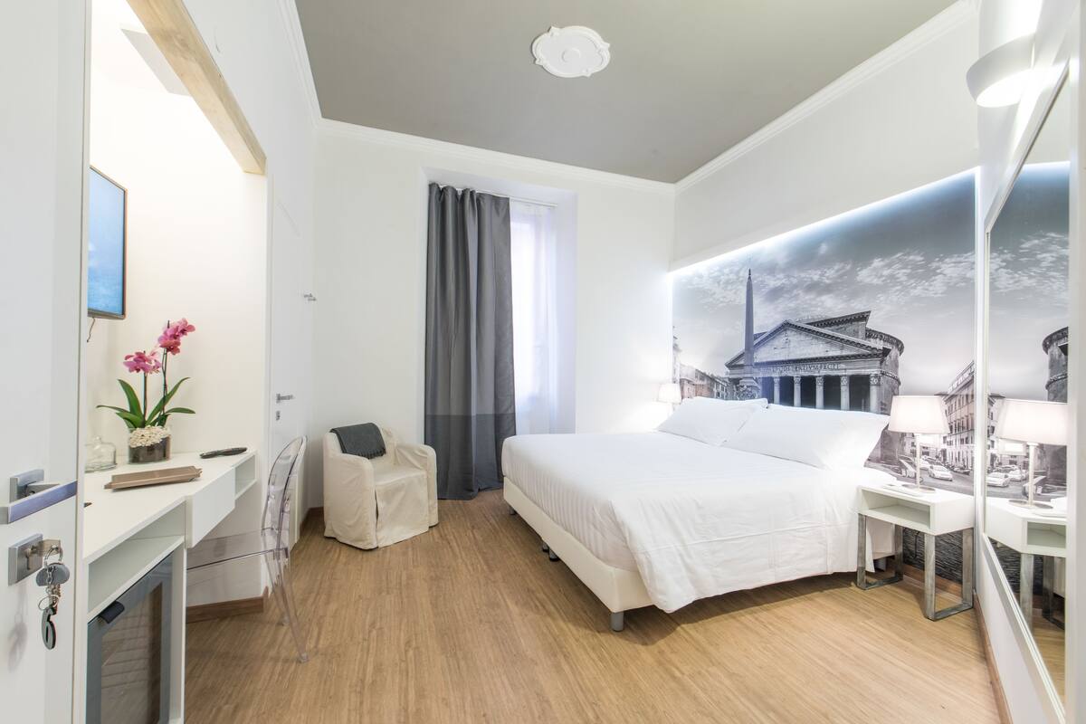 A modern hotel room with a large bed, a desk, a chair, a small table with a lamp, and a mirror.